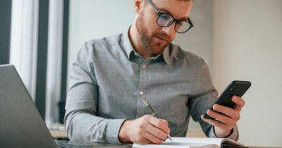Ein Mann prüft Informationen auf dem Smartphone und macht Notizen am Schreibtisch. Das Bild steht für die Dokumentation geschäftlicher Fahrzeugnutzung. Fleethouse erklärt die 1-Prozent-Regelung verständlich und unterstützt Unternehmen mit digitalem Gewerbeleasing und transparenter Fuhrparkverwaltung.