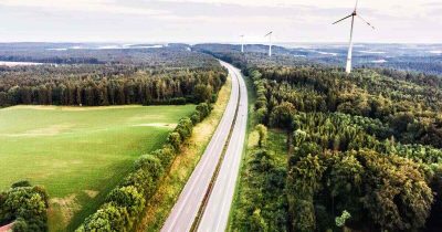 Das Bild zeigt eine weitl&auml;ufige Stra&szlig;e, ges&auml;umt von B&auml;umen und Windr&auml;dern, die die Nutzung erneuerbarer Energien symbolisieren. F&uuml;r Unternehmen bietet Fleethouse L&ouml;sungen f&uuml;r Fuhrparks mit alternativen Antrieben, um nachhaltige Mobilit&auml;t zu f&ouml;rdern. So k&ouml;nnen Sie Ihre Flotte zukunftsf&auml;hig und umweltbewusst gestalten.