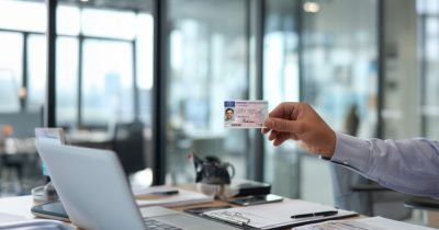 Ein Fuhrparkleiter h&auml;lt einen F&uuml;hrerschein zur Kontrolle in der Hand. Vor ihm ist ein Laptop und eine Checkliste. Ein Sinnbild f&uuml;r die F&uuml;hrerscheinkontrolle Arbeitgeber Muster